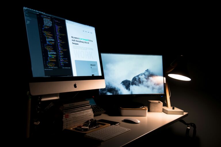 Dual computer monitors sit on a dark desk; one shows code, the other a mountain image. A lit lamp casts a warm glow, forming a focused, peaceful workspace.
